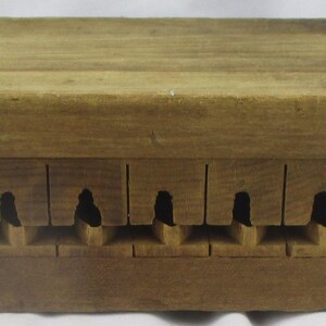 May include: A rectangular wooden mold featuring a series of twelve evenly spaced vertical slots along the side, each containing a small carved cavity. The natural brown wood grain is visible across the entire surface of this functional tool.