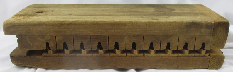 May include: A rectangular wooden mold featuring a series of twelve evenly spaced vertical slots along the side, each containing a small carved cavity. The natural brown wood grain is visible across the entire surface of this functional tool.
