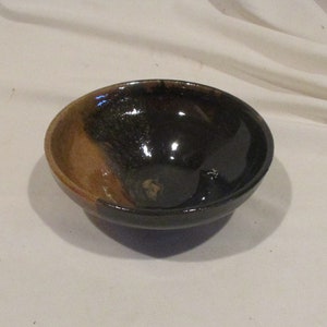 May include: A small, round ceramic bowl with a black and brown glaze. The bowl has a glossy finish and is slightly uneven in shape.