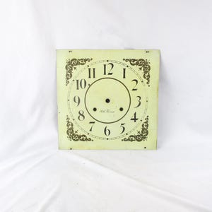 May include: A square clock face with a pale yellow background and black numerals. The numbers 1 through 12 are displayed, with decorative flourishes in each corner. The words "Seth Thomas" are printed near the center.