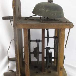 May include: An antique wooden clock mechanism with a bell on top. The clock has visible gears and a wooden frame. The bell is dark green and sits on a wooden platform. The clock's wooden frame is a light brown color.