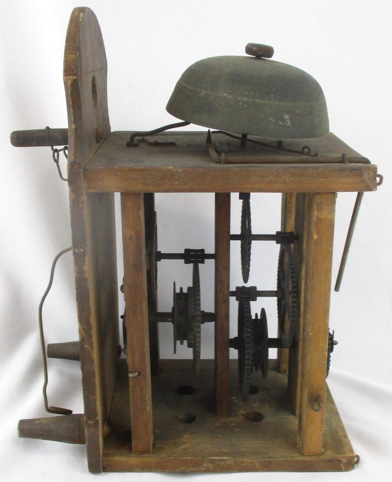 May include: An antique wooden clock mechanism with a bell on top. The clock has visible gears and a wooden frame. The bell is dark green and sits on a wooden platform. The clock's wooden frame is a light brown color.