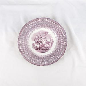 May include: A round, decorative plate with a scalloped edge. The plate features a detailed, repeating pattern in a mauve color around the rim. The center of the plate has a mauve illustration of figures and a dog near a decorative urn.
