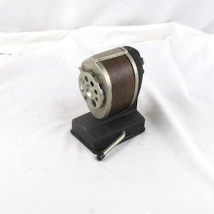 May include: A vintage pencil sharpener with a black base, a silver metal blade housing, and a brown wooden barrel. The sharpener has a silver crank handle and a classic design.