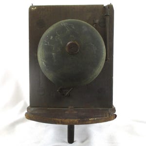 May include: Antique wall-mounted bell with a dark, weathered appearance. The bell is a large, rounded shape, mounted on a rectangular wooden backboard. A small wooden shelf is below the bell. The bell has a dark, aged patina, suggesting it is vintage.