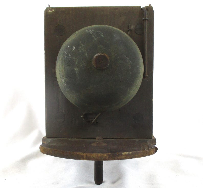 May include: Antique wall-mounted bell with a dark, weathered appearance. The bell is a large, rounded shape, mounted on a rectangular wooden backboard. A small wooden shelf is below the bell. The bell has a dark, aged patina, suggesting it is vintage.
