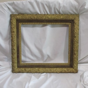 May include: A gold-colored picture frame with ornate detailing. The frame has a dark brown inner edge and a raised, textured outer edge.
