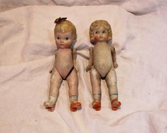 May include: Two vintage porcelain dolls with painted details. The dolls are nude with blue and orange painted accents on their legs and feet. The dolls have blonde hair and painted facial features.