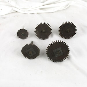 May include: Five antique metal gears of varying sizes are arranged on a white surface. The gears have a dark, aged appearance with visible teeth. The gears appear to be from a clock or other mechanical device.