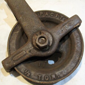May include: A vintage cast iron pulley with a 1/2 ton capacity. The pulley has a large wheel with a groove for a rope and a lever arm with a nut and bolt. The pulley is rusty and has a worn surface.