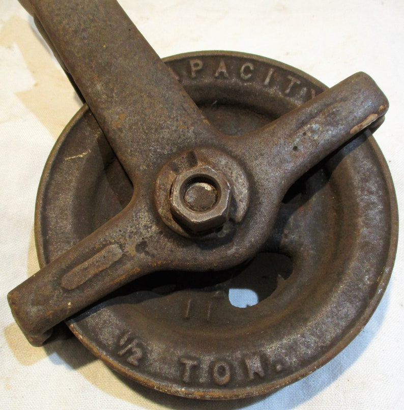 May include: A vintage cast iron pulley with a 1/2 ton capacity. The pulley has a large wheel with a groove for a rope and a lever arm with a nut and bolt. The pulley is rusty and has a worn surface.