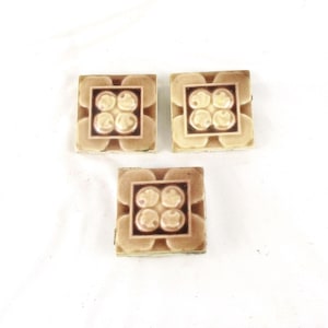 May include: Three square ceramic tiles with a raised design of four stylized flowers in a light brown color with a darker brown border.