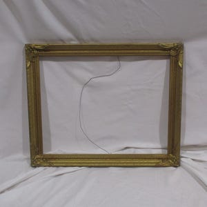 May include: A gold picture frame with ornate detailing. The frame is empty and has a wire hanging from the top.