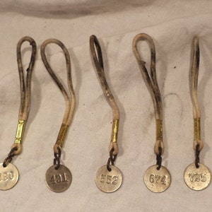May include: Five antique metal tags with leather straps. The tags are round and have the numbers 330, 421, 552, 674, and 725 printed on them.