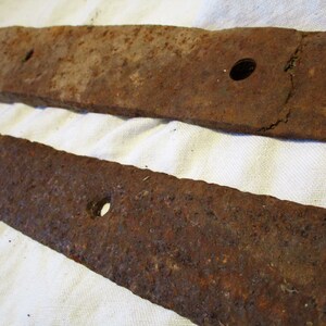 May include: Two rusty metal hinge straps with holes for attaching to a door or gate. The straps are made of a dark brown metal with a rough texture.