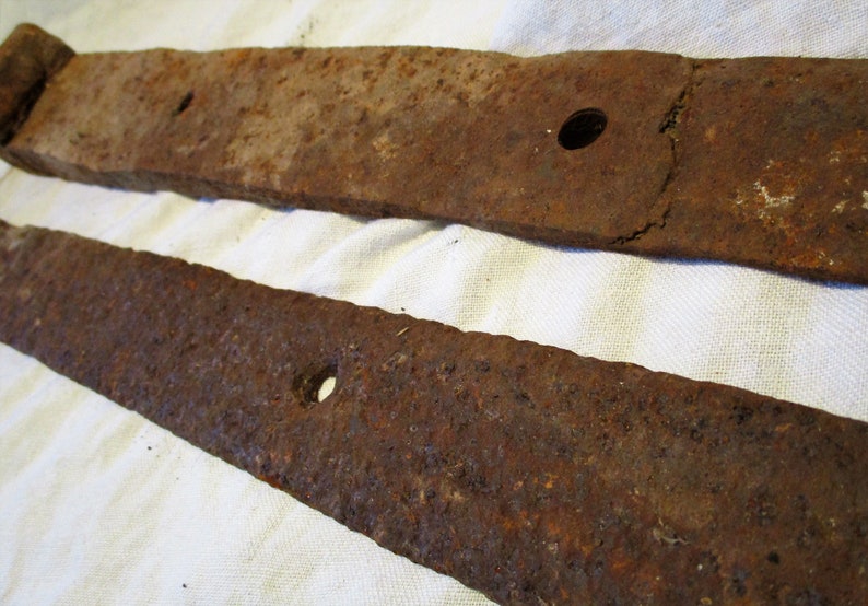 May include: Two rusty metal hinge straps with holes for attaching to a door or gate. The straps are made of a dark brown metal with a rough texture.
