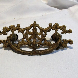 May include: A gold-toned metal drawer pull with a decorative, ornate design. The pull has a curved shape and two decorative knobs on either side.