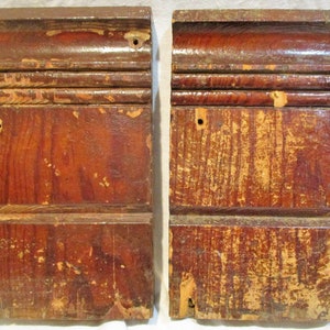 May include: Two antique wooden corbels with a reddish-brown finish and a decorative carved design. The corbels have a distressed look with some paint chipping and wear.