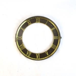 May include: A circular clock face with Roman numerals. The face is dark brown with gold-colored numerals and a gold-colored outer rim. The clock face is a vintage or antique item.