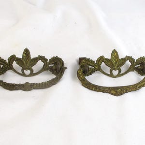 May include: Two antique brass drawer pulls with a decorative leaf and scroll design. The pulls have a curved handle and a central leaf motif. The brass has a tarnished, aged appearance, suggesting vintage or antique status.