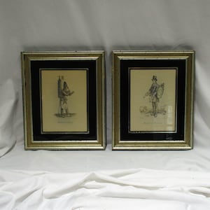 May include: Two framed black and white drawings of men in historical attire. The drawings are set against a cream background and are framed in gold with a black mat.