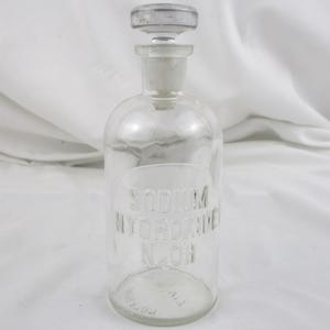 Może przedstawiać: Przezroczysta szklana butelka z korkiem. Butelka ma zaokrąglony korpus i długą szyjkę. Na przodzie wytłoczono napisy "SODIUM HYDROXIDE N.OH". Korek jest przezroczysty i ma zaokrągloną g&oacute;rę.