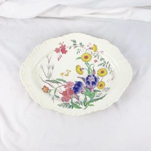 May include: An oval-shaped, off-white serving platter with a scalloped edge. The platter is decorated with a floral design featuring yellow, red, blue, and purple flowers with green leaves. The flowers are arranged in a cluster.