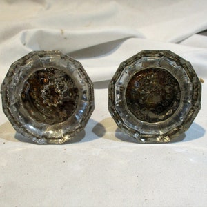 May include: Two antique glass door knobs with a faceted design and a clear glass finish. The knobs have a decorative pattern on the front.