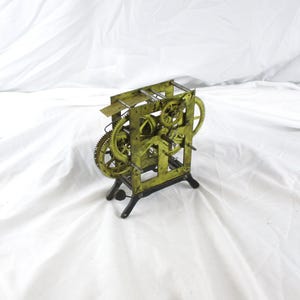 May include: A brass clock movement mechanism featuring an exposed gear train with multiple interlocking wheels and pinions mounted within a rectangular frame, supported by a dark metal base with four feet, showcasing intricate mechanical parts.