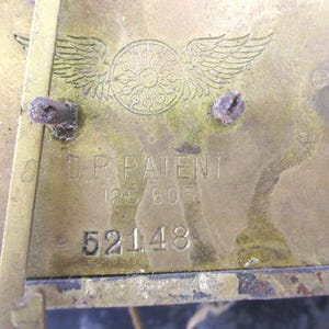 Peut inclure: Gros plan d'une plaque en laiton avec le texte "D.P. PATENT" et les chiffres "125 605" et "52148". La plaque pr&eacute;sente un embl&egrave;me d&eacute;coratif avec des ailes et un motif circulaire. Le laiton a un aspect terni.