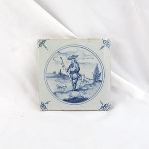 May include: Square ceramic tile with a blue and white pastoral scene. The image depicts a man with a staff, sheep, and trees within a circular frame. Decorative blue corner accents complete the design.