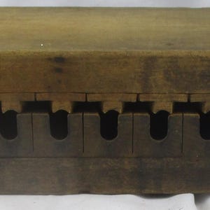 May include: A rectangular brown wooden mold featuring two interlocking sections. The top piece has a series of protruding wooden tabs, while the bottom piece contains a corresponding row of ten U-shaped cutouts along its length.