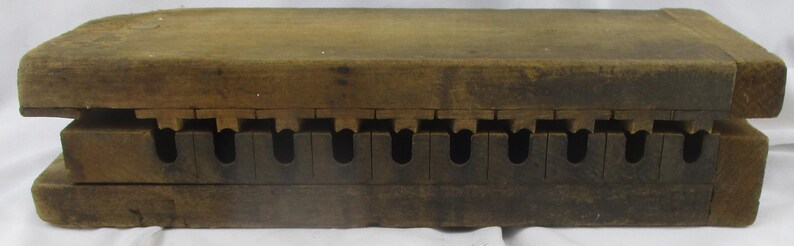 May include: A rectangular brown wooden mold featuring two interlocking sections. The top piece has a series of protruding wooden tabs, while the bottom piece contains a corresponding row of ten U-shaped cutouts along its length.