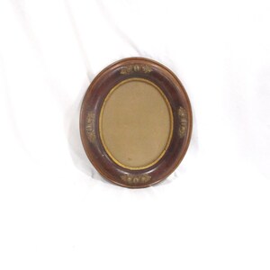 May include: Oval-shaped, dark brown picture frame with a gold inner border. The frame has decorative gold accents on the outer edge. The frame is empty, with a tan backing visible inside.
