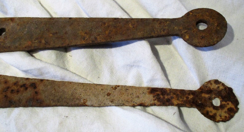 May include: Two rusty metal hinges with rounded ends and a hole in the center. The hinges are on a white fabric background.