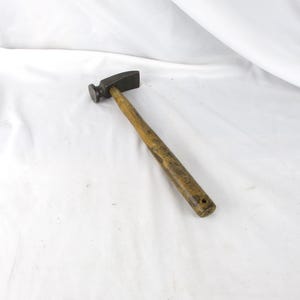 May include: A small, antique hammer with a dark metal head and a worn wooden handle. The hammer head is rectangular and round, and the handle is a light brown color. The tool is designed for precision work.