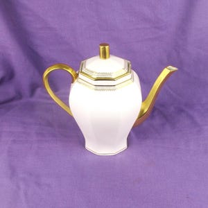 May include: White porcelain teapot with a gold lid and handle. The teapot has a geometric design and is decorated with gold trim.