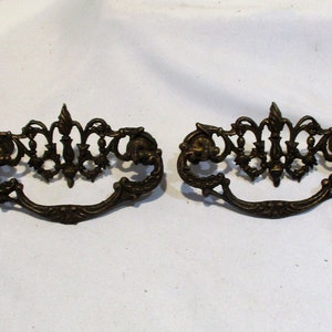 May include: Two ornate brass drawer pulls with a decorative, scroll-like design. The pulls have a slightly aged, antique finish.