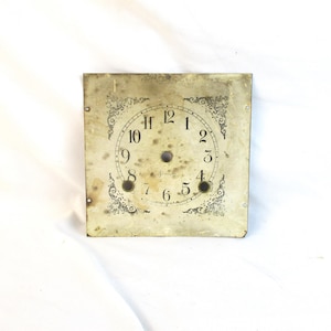 May include: A square, antique clock face with black numerals and ornate corner designs. The face is aged with visible wear and tear, including spots and discoloration. The clock face has a central hole and smaller holes for clock hands.