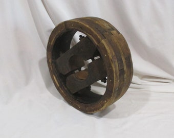 May include: A large wooden pulley with a thick, layered rim and a central hub with a complex, interlocking design. The pulley is made of dark brown wood and has a rustic, antique appearance.