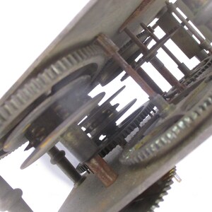May include: Close-up of a vintage clock mechanism. The image shows a complex arrangement of metal gears, rods, and other components. The gears are various sizes and have a worn, aged appearance, suggesting the clock's age. The overall color is a mix of bronze and rust.