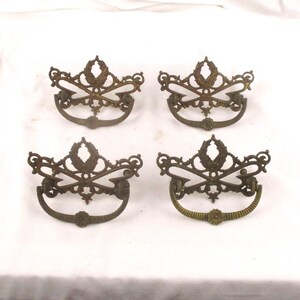 May include: Four antique brass drawer pulls with ornate designs. Each pull features a central floral motif with intricate scrollwork and a curved handle.