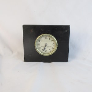 May include: A small, vintage clock with a round face set in a black rectangular block. The clock face has black numerals and hands, with a gold-colored bezel. The clock is likely an antique desk or shelf clock.