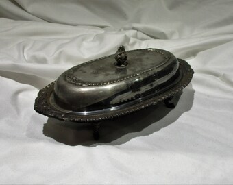 May include: A silver oval butter dish with a glass lid and a decorative knob on top. The dish has a beaded edge and sits on three feet.