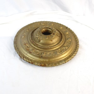 May include: A circular, metallic gold-toned ceiling canopy or light fixture plate featuring a raised rope-style border, embossed floral patterns, and a central threaded opening for a light socket, showing signs of age and patina.