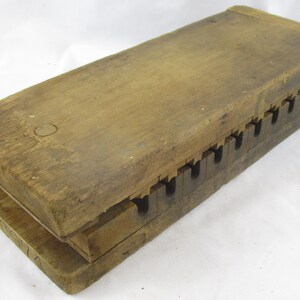 May include: A rectangular wooden press featuring two thick, weathered planks joined by a series of interlocking, notched teeth along one side. The wood displays a natural, aged brown grain with visible surface texture and a simple, functional construction.