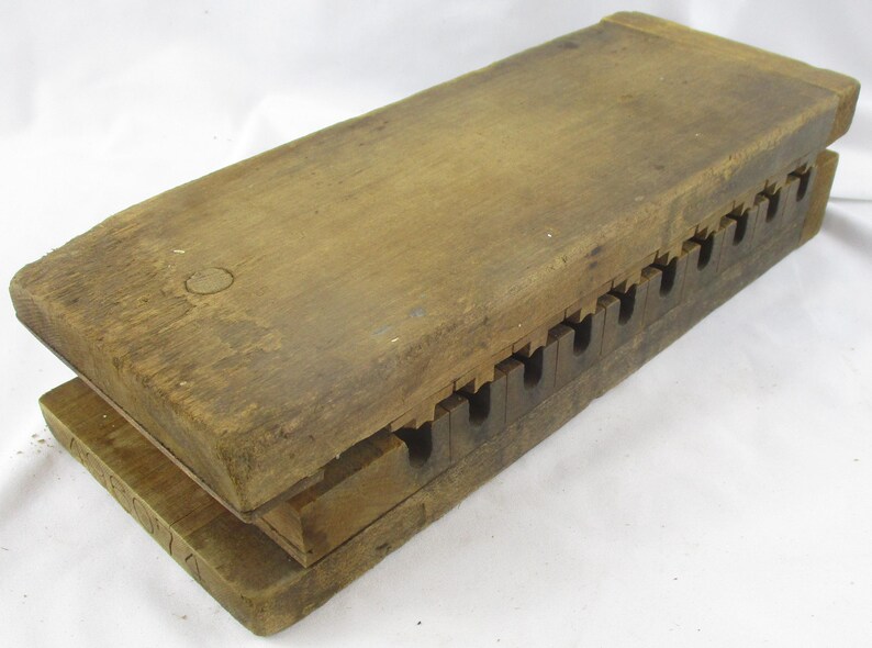 May include: A rectangular wooden press featuring two thick, weathered planks joined by a series of interlocking, notched teeth along one side. The wood displays a natural, aged brown grain with visible surface texture and a simple, functional construction.