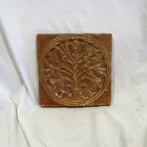May include: A square ceramic tile with a raised design of a stylized floral motif in a circle. The tile is brown with a yellow glaze.