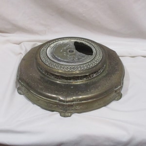 May include: A round, gold-toned metal base with a decorative design and a center hole. The base has a raised edge and a scalloped design around the bottom.