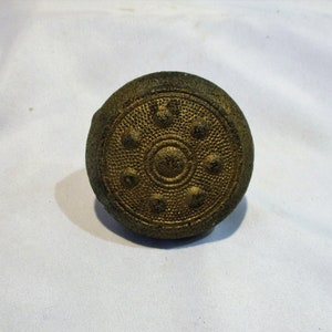 May include: A round, gold-colored metal button with a raised, circular design and six smaller circles around the center.